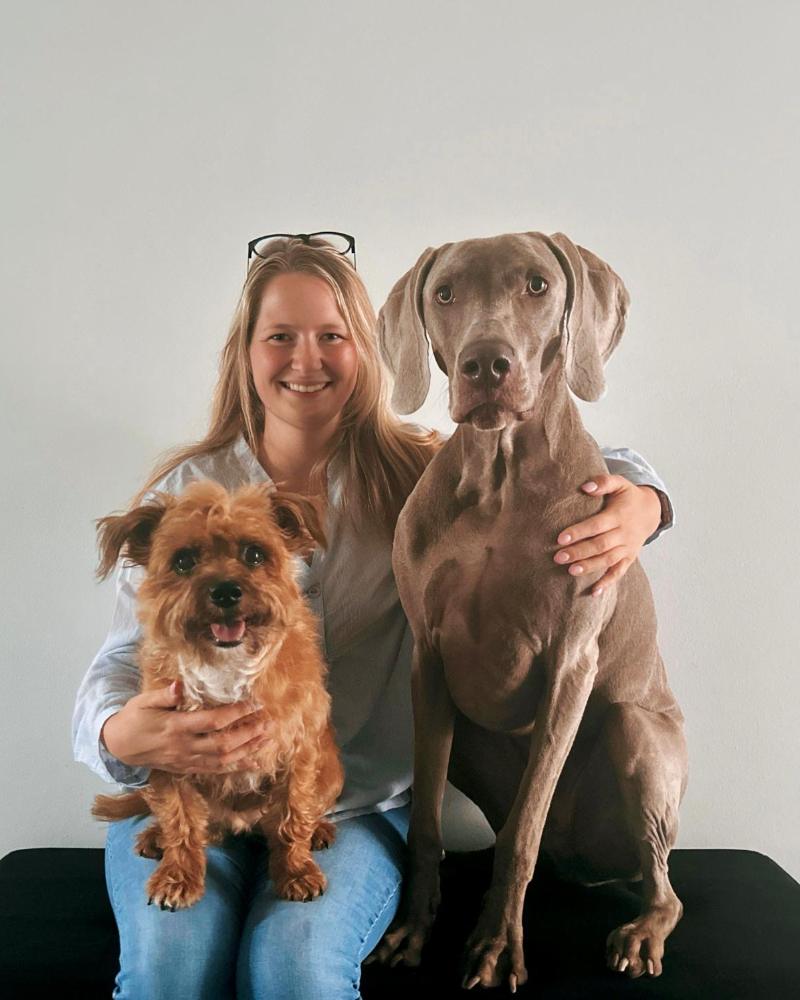 Corinna sitzt zwischen ihren beiden Hunden – links ein kleiner rotbrauner Mischling auf ihrem Schoß, rechts ein Weimaraner.